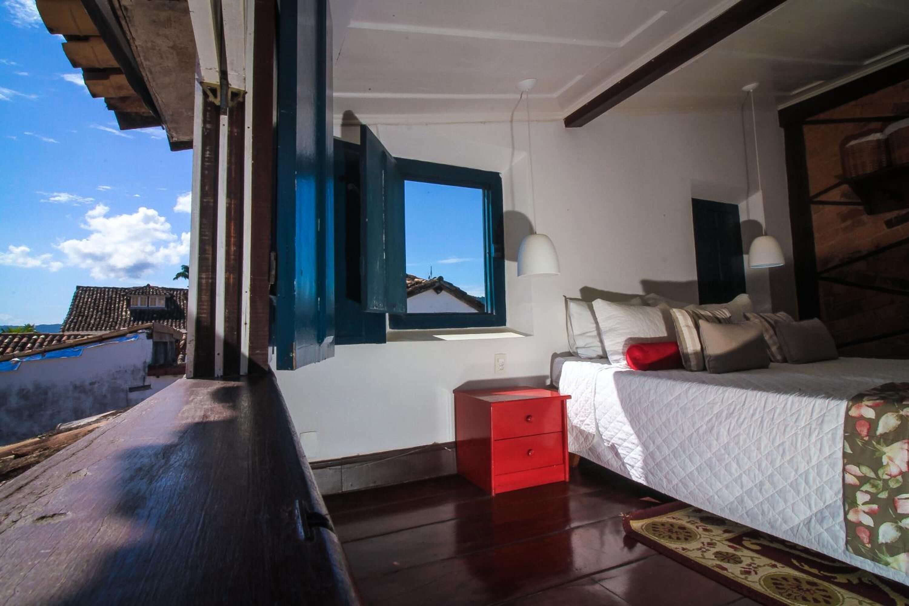 Un dormitorio con una cama, una mesita de noche roja, una ventana con contraventanas azules y vistas al exterior.
