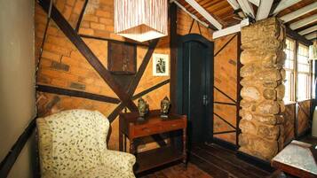 A rustic room with exposed wooden beams, a stone wall, a patterned armchair, a wooden table with decorative items, and a dark door.