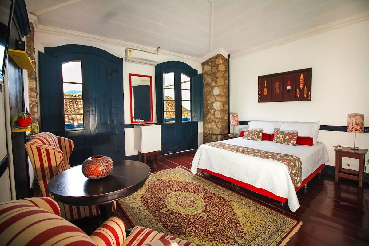 Quarto de hotel com uma cama grande, uma mesa redonda com um vaso, cadeiras listradas e vista através de uma porta.