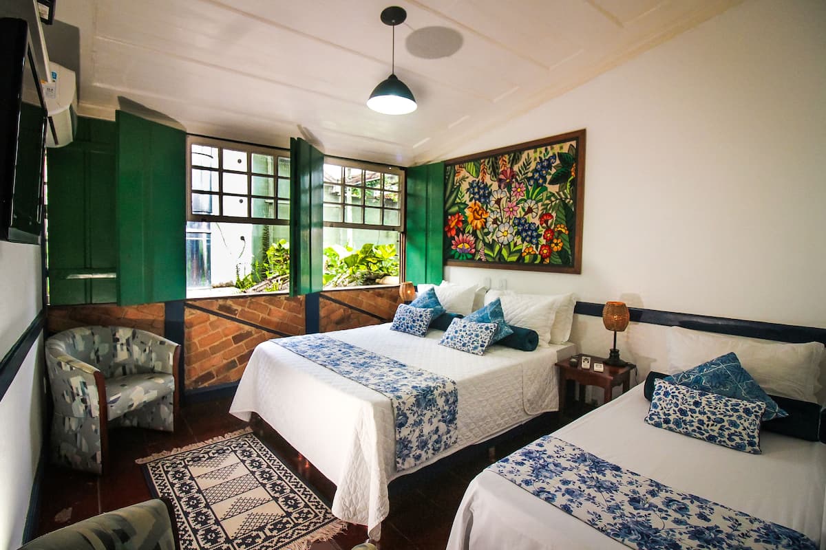 Quarto de hotel com duas camas, uma cadeira verde, uma mesa pequena e janela com vista para vegetação.