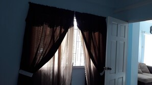A room with a door leading to a bright area, a window with curtains, and a sofa in the background. - Corinth Grove Apartment (St. David)