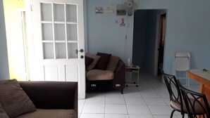 A living room with a brown sofa, a white door, a small table with a fan, and a tiled floor. - Corinth Grove Apartment (St. David)