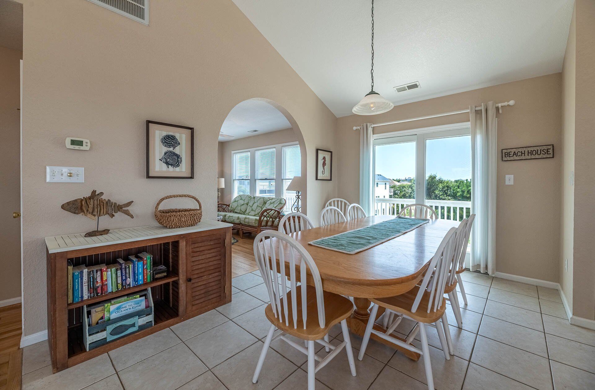 A dining area with a wooden table and white chairs, a bookshelf with books, a framed fish skeleton, and a sign reading "BEACH HOUSE".