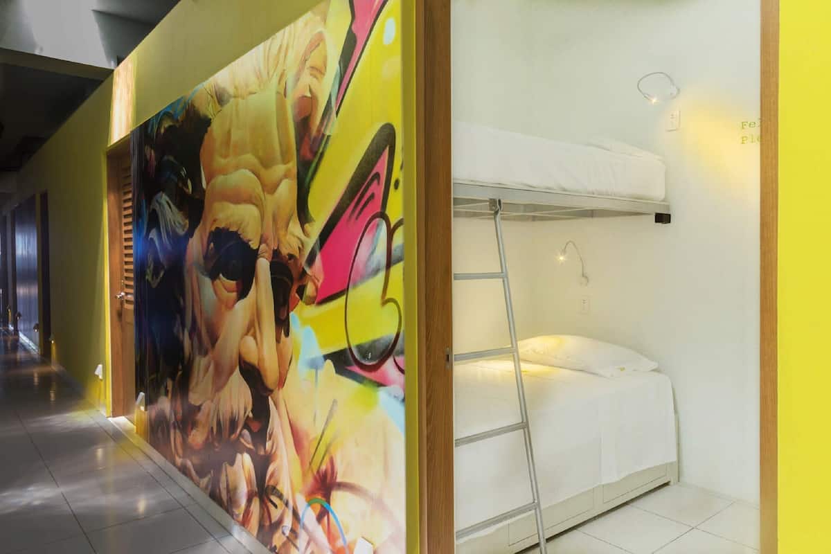 Una habitación de hotel con literas, un mural de colores en la pared y una mesita de noche pequeña con lámpara.