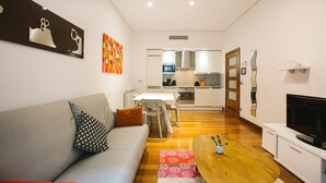 Una sala de estar moderna con un sofá gris, una mesa de centro de madera y un televisor. Se ve una cocina al fondo con armarios blancos y un microondas.