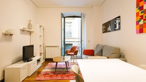 Un dormitorio moderno y bien iluminado con cama, televisor, una mesita y una silla. Se ve una ventana con vistas a un balcón.