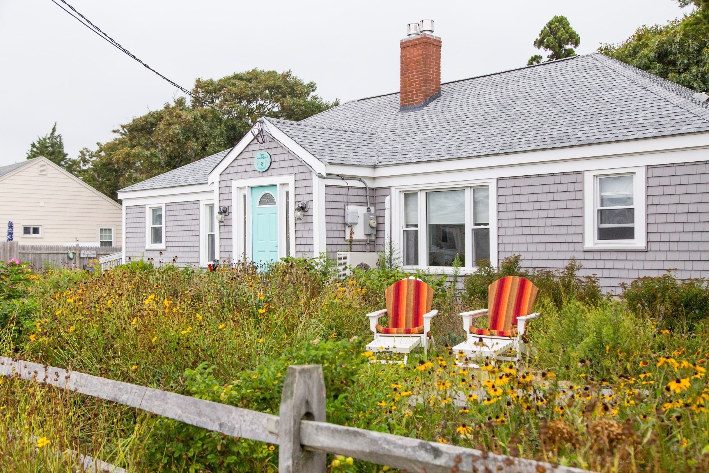 A house with a grey exterior and a blue door, surrounded by a garden with various plants and flowers.