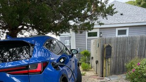 Ein blaues Auto steht auf einem Schotterweg mit einem Holzzaun und einem Haus im Hintergrund.