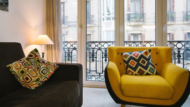 Een kamer met een donkere bank en een gele fauteuil, beide met gekleurde kussens, en uitzicht op een balkon met sierhekwerk.