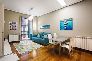 A modern living room with a teal sofa, a dining table with chairs, a patterned rug, and artwork on the walls.
