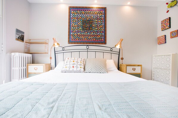 A neatly arranged bedroom with a bed, bedside tables, a radiator, and a wall with geometric artwork.