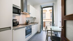A modern kitchen with white cabinets, a built-in oven, and a coffee maker. There is a dining area with a table and chairs, and a view of a balcony with a railing. - 4 Bella Easo B San Marcial 28 (San Sebastián)