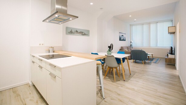 Una cocina moderna con una isla central, un área de comedor con mesa y sillas, y una sala de estar con sofá y televisor.