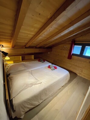 Een kamer met een houten plafond, twee bedden met witte lakens, een rood gordijn en een raam met zonwering.