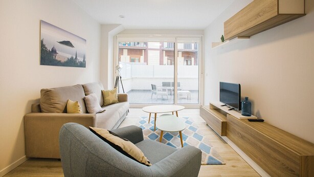 A modern living room with a sofa, a coffee table, a TV, and a balcony with a view of buildings. - M Gloria Suites 5 1C  inner courtyard, one bedroom (San Sebastián)