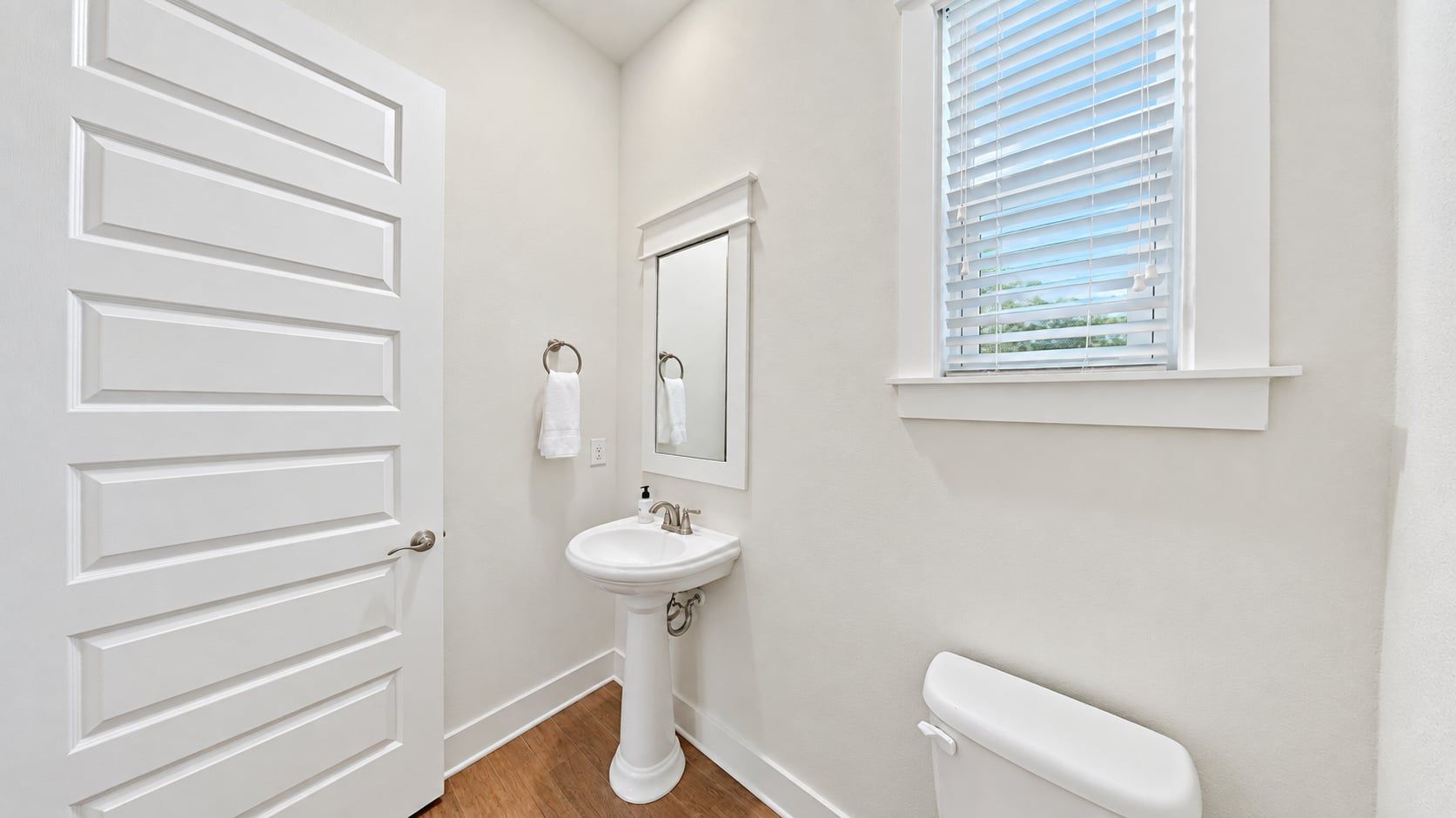 Une salle de bain avec une porte blanche, un lavabo sur colonne, des toilettes et une fenêtre avec des stores.