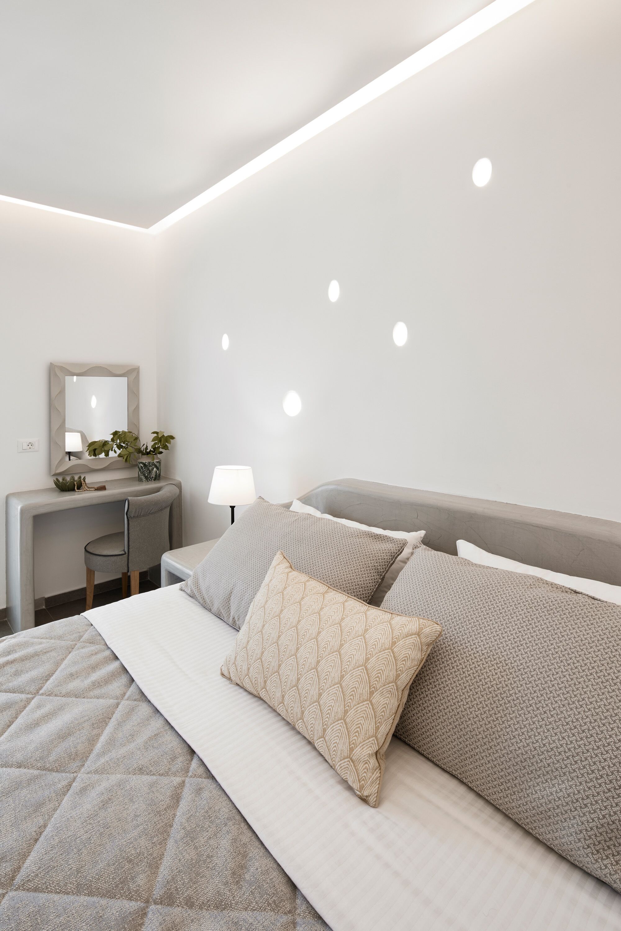 A modern bedroom with a bed, bedside table, lamp, and a desk with a mirror and plants.
