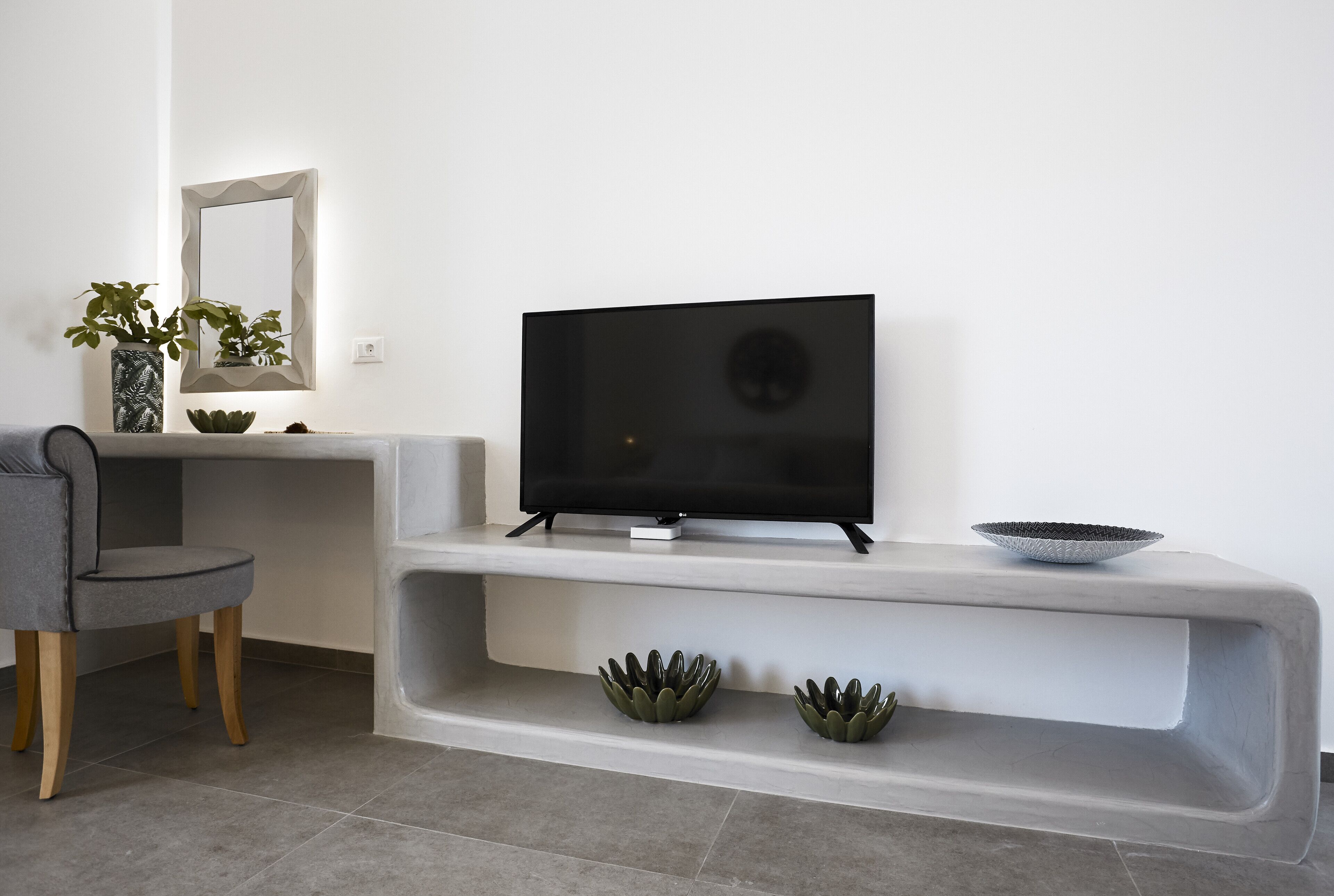Een moderne woonkamer met een flatscreen televisie op een witte console, een grijze stoel en decoratieve plantenpotten.