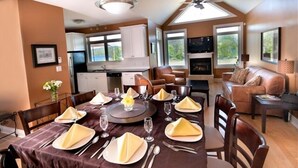 Comedor con una mesa dispuesta para cuatro, cocina al fondo y sala de estar con chimenea y asientos.