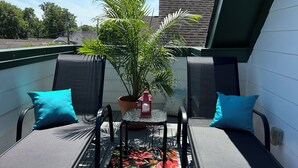 A balcony with two black metal chairs, each with a teal pillow, a small table with a red object, and a colorful floral rug.