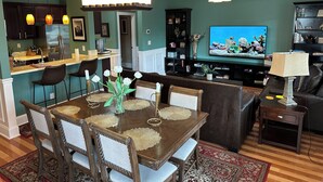 A modern kitchen with a central island, a dining area with a wooden table and chairs, a living room with a sofa and a TV displaying an underwater scene, and a ceiling fan.