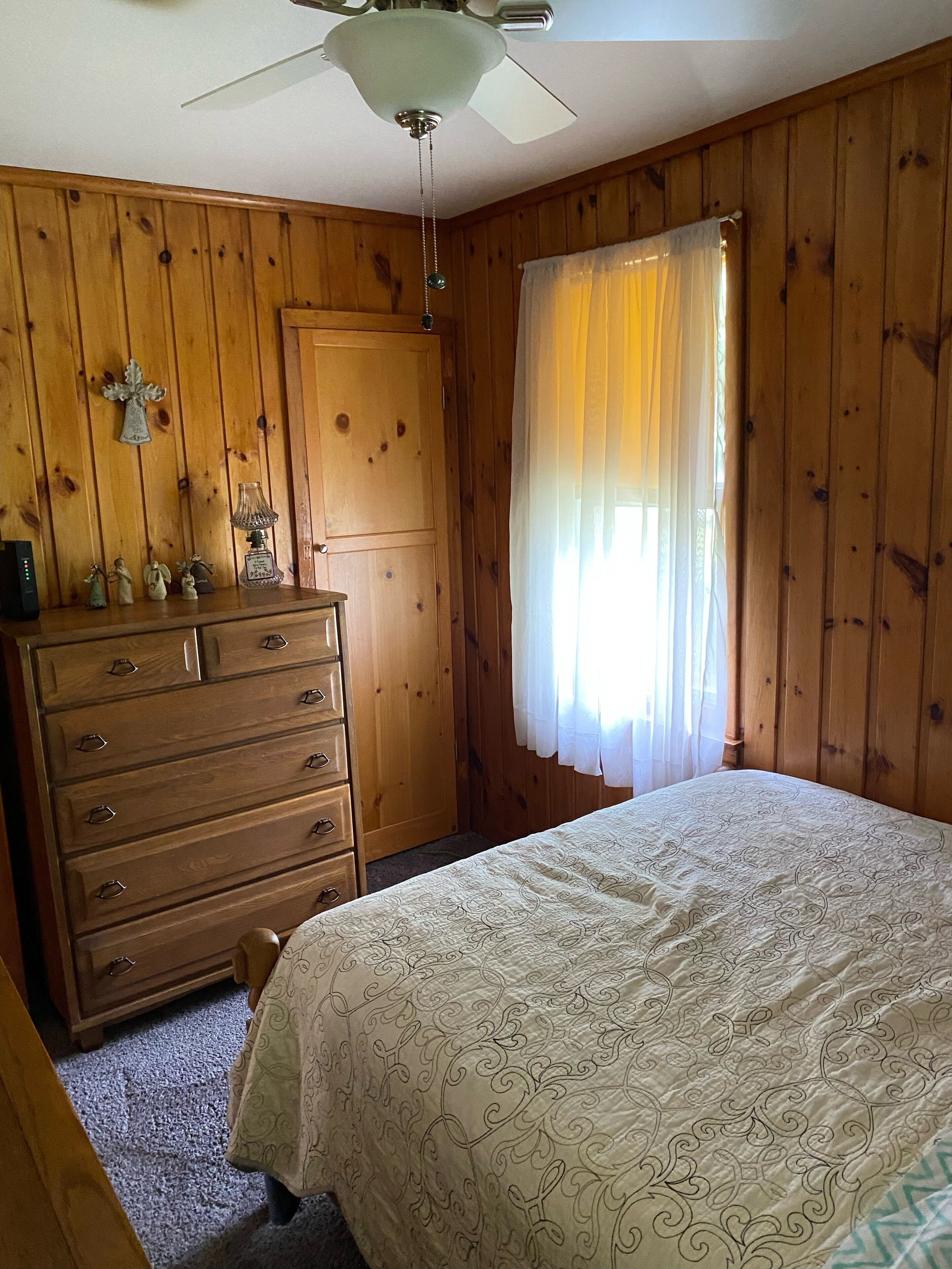 A bedroom with wooden paneling, a bed with a patterned bedspread, a dresser with drawers, a ceiling fan, and a window with sheer curtains.