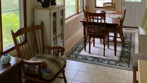 A dining area with a wooden table and chairs, a cabinet with decorative items, and large windows with a view of greenery.