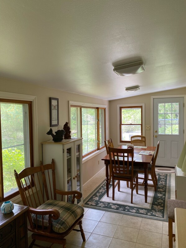 A dining area with a wooden table and chairs, a cabinet with decorative items, and large windows with a view of greenery.