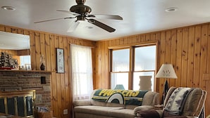 A cozy living room with wooden walls, a ceiling fan, a fireplace, a sofa, a coffee table, and a lamp.