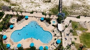 An aerial view of a resort with a large central pool, multiple sun loungers, and a surrounding sandy area.
