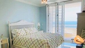 A bedroom with a bed, a nightstand, a glass table, and a view of the sea through open shutters.