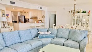 A living room with a light blue sectional sofa, a small white coffee table, and a kitchen in the background.