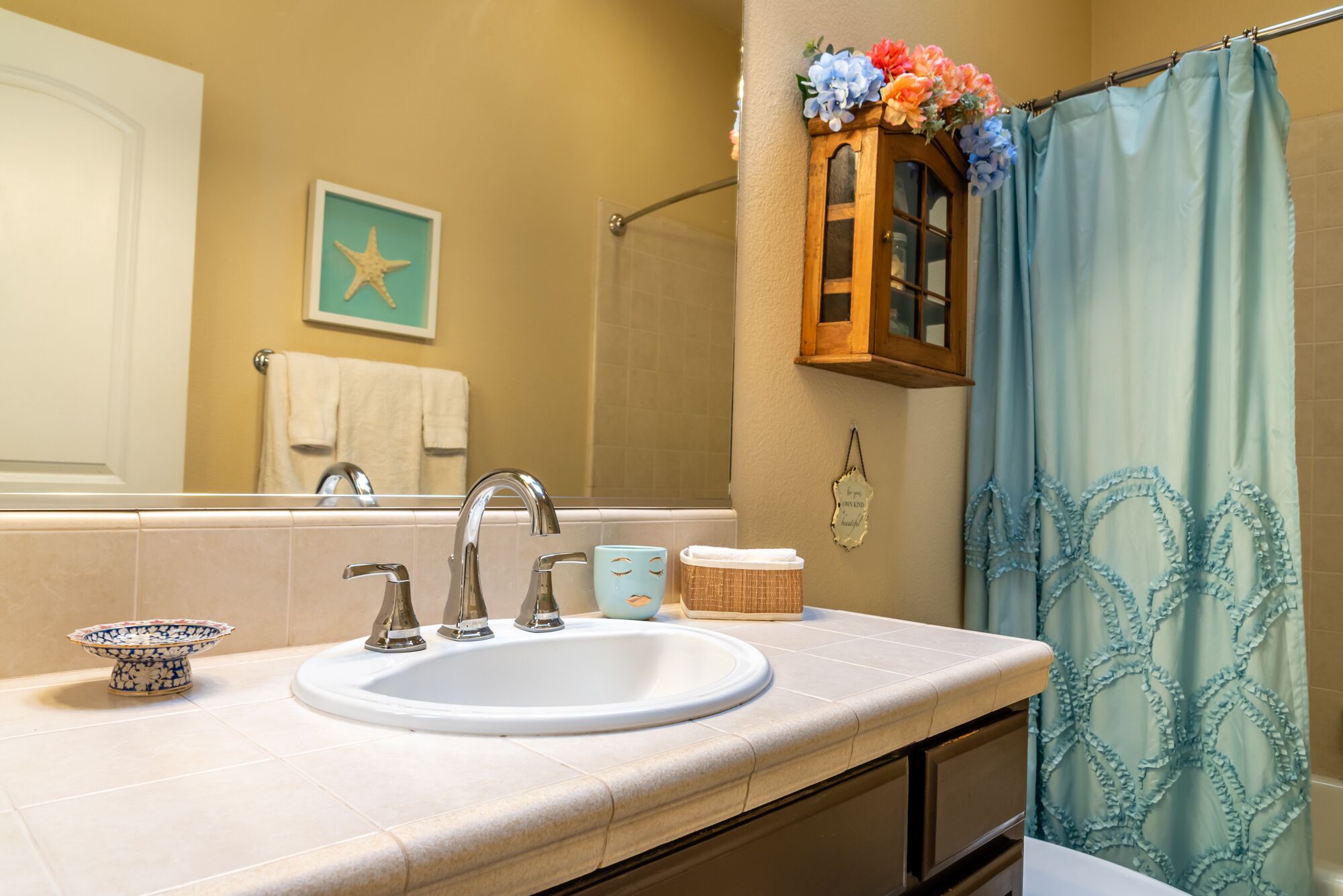 Une salle de bain comprenant un lavabo, deux robinets, un miroir, un porte-serviettes, une étoile décorative, un meuble en bois orné de fleurs, un panier et un rideau de douche à volants.