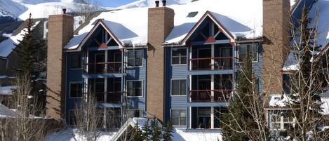 A multi-story hotel with a ski resort backdrop, featuring a prominent chimney, large windows, and a balcony on the second floor.