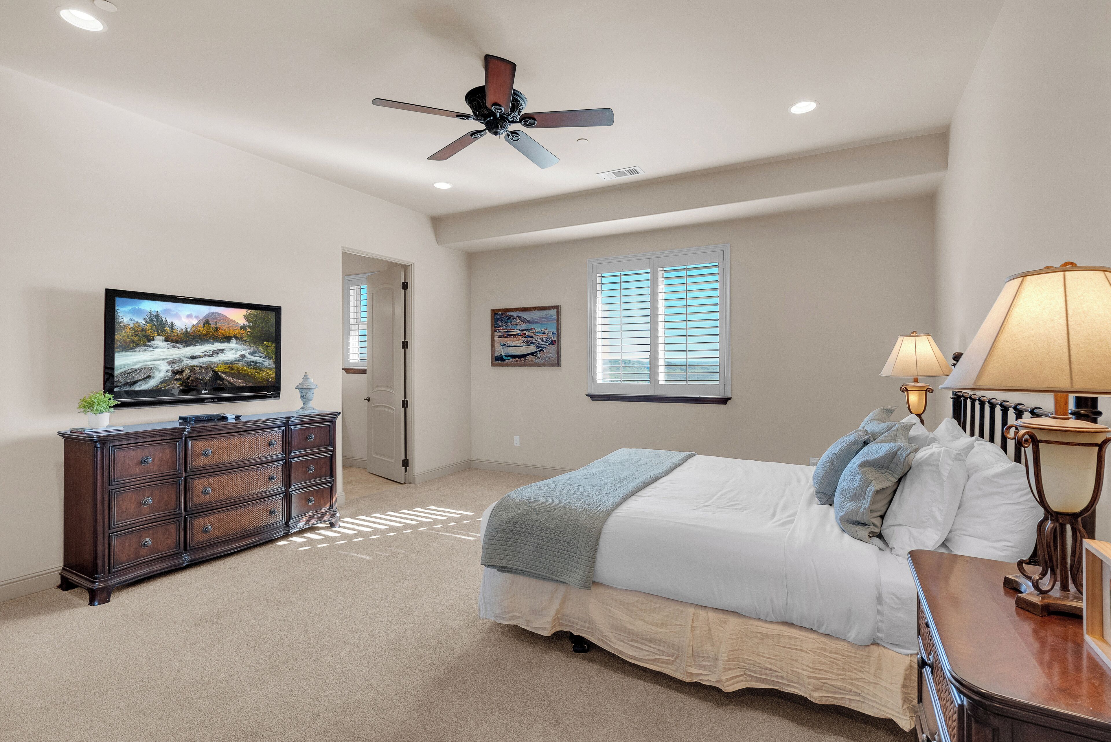 A bedroom with a bed, dresser, TV, ceiling fan, window, and lamp.
