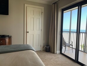 A bedroom with a bed, a dresser, a door, a trash can, and a balcony with a view of the ocean.