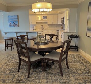 A dining area with a round table, six chairs, a chandelier, and a kitchen in the background.