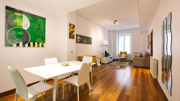 A modern living room with a white dining table, white chairs, a sofa, and a TV.