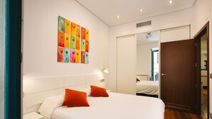 A hotel room with a large bed, two orange pillows, a white headboard, a bedside table, a nightstand, a wall-mounted lamp, a mirror, and a door.
