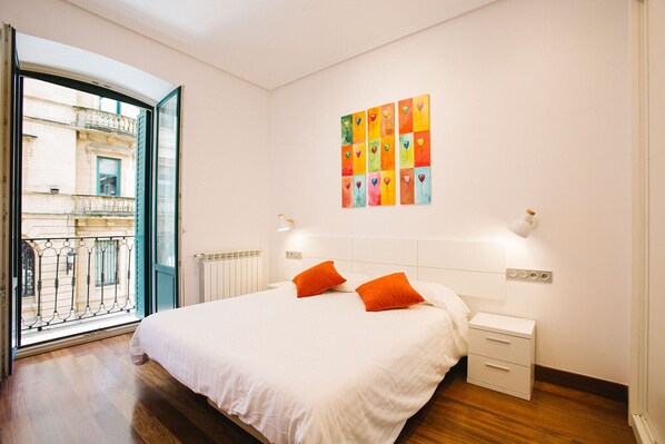 A bedroom with a bed, two nightstands, a radiator, a window with a view of a building, and a colorful wall art piece.