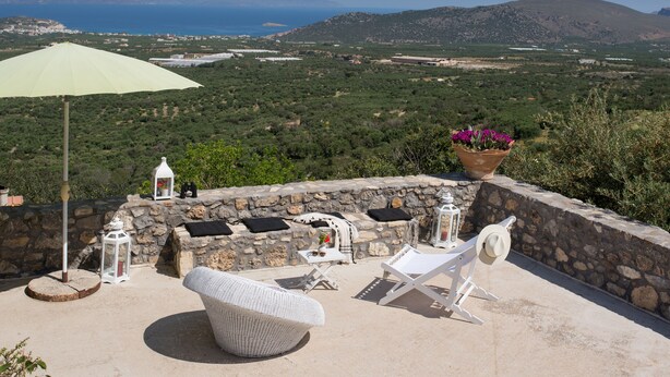 A terrace with a view of the sea and mountains, stone walls, a wicker chair, a table with a lantern, and a potted plant. - ELIA village house . (Monastiraki ierapetra)