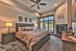 A bedroom with a large bed, a ceiling fan, a fireplace, and a view of the outdoors.