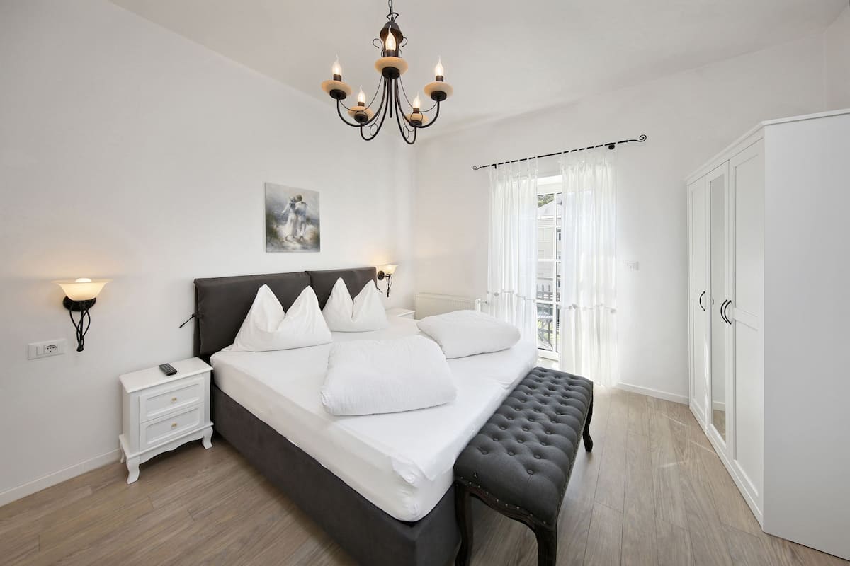 A bedroom with a large bed, bedside lamps, a chandelier, a bench, and a wardrobe.