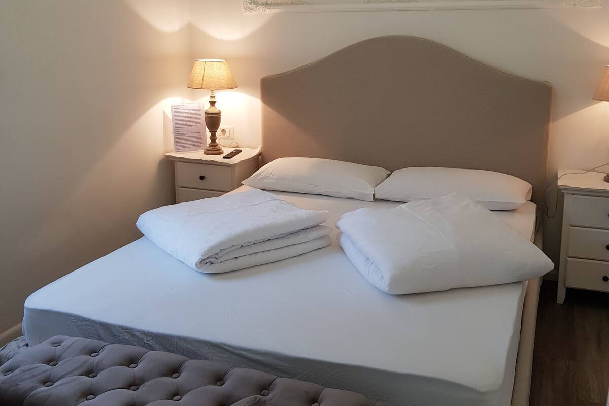 A neatly made bed with white linens, a tufted bench, and a nightstand with a lamp and a remote control.