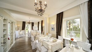 A formal dining room with multiple tables set for service, a chandelier, and a view of the outdoors through a window. - Garni Villa Tyrol - Adults Only (Merano)