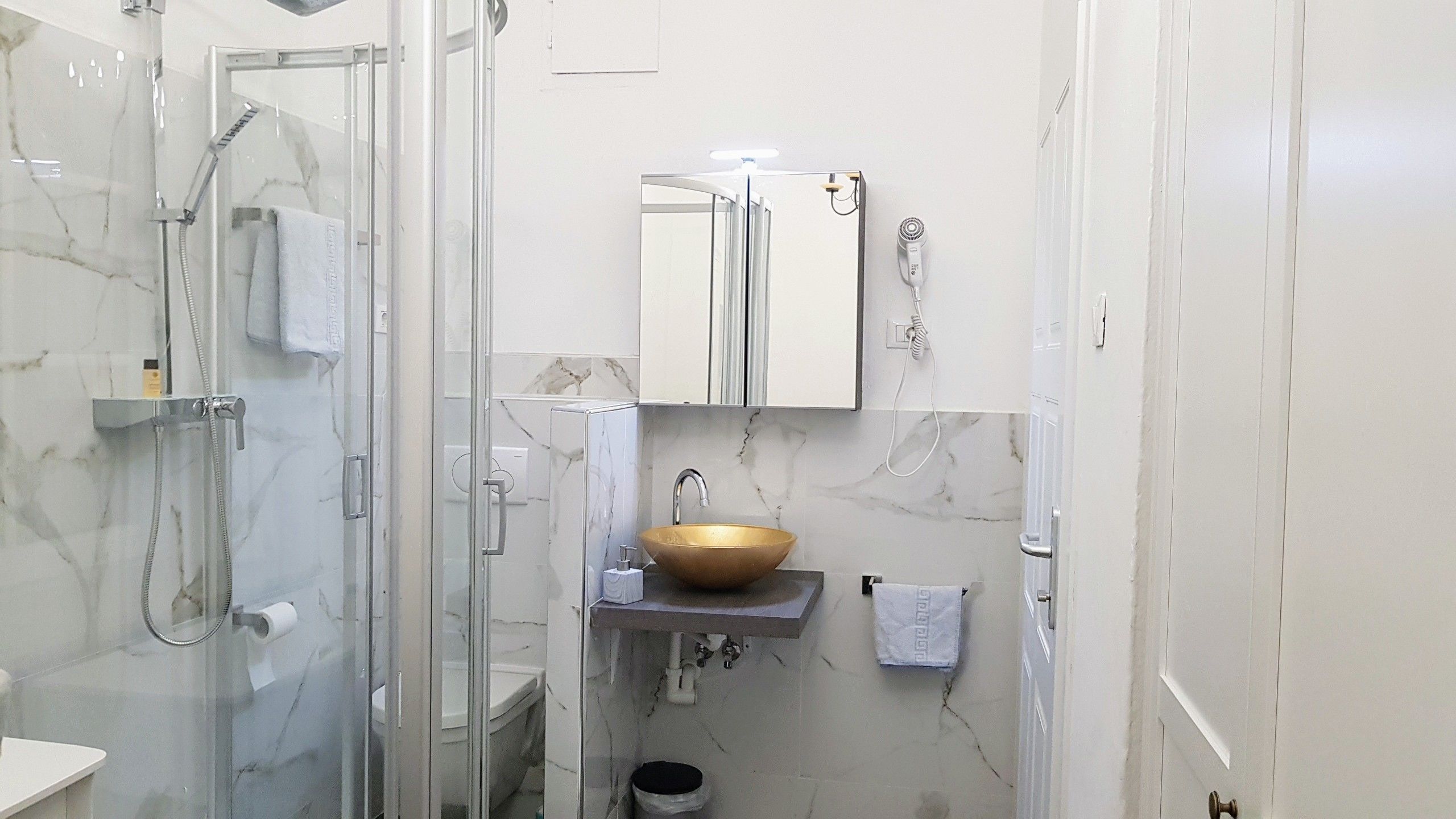 Une salle de bain dotée d’une douche à parois de verre, d’un mur recouvert de carreaux de marbre, d’un lavabo sur pied doré et de toilettes blanches.