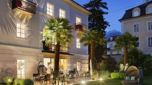 A well-lit outdoor seating area with tables and chairs, surrounded by palm trees and potted plants. - Garni Villa Tyrol - Adults Only (Merano)