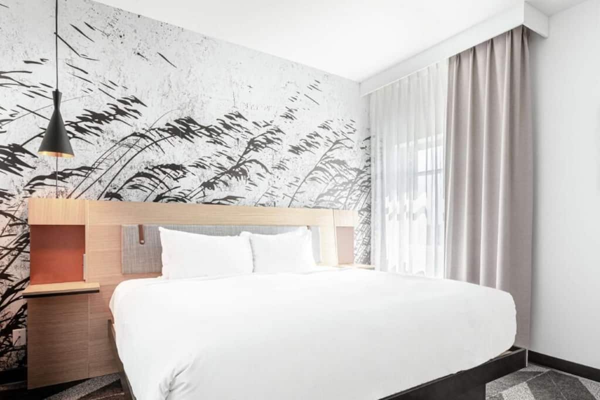 Et moderne hotelværelse med en stor seng, et træhovedgærde, et natbord, en hængende lampe og en væg med bladmotiv.
