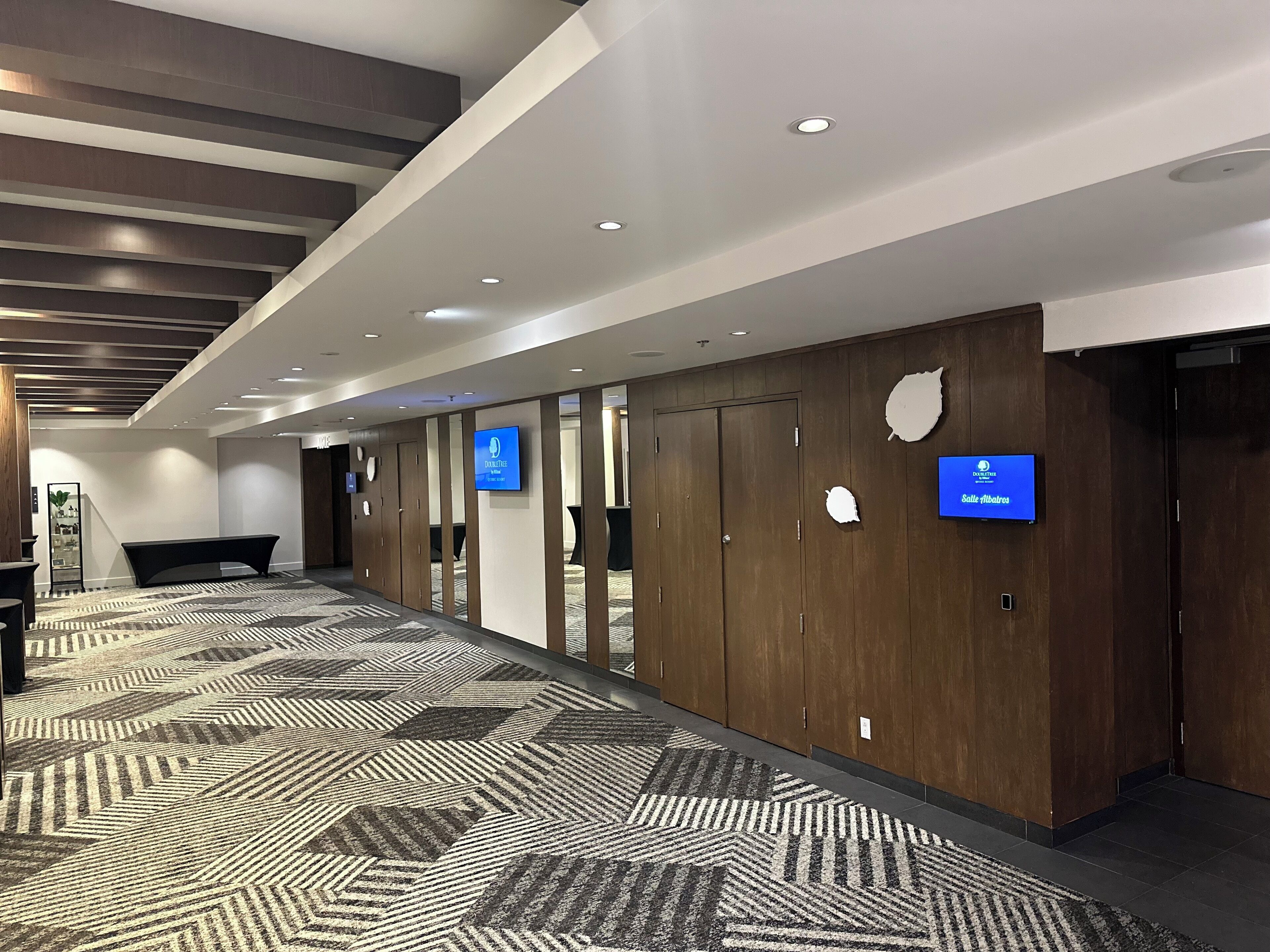 A modern hotel lobby with a patterned carpet, wooden panel walls, and a flat-screen TV displaying the hotel's name.
