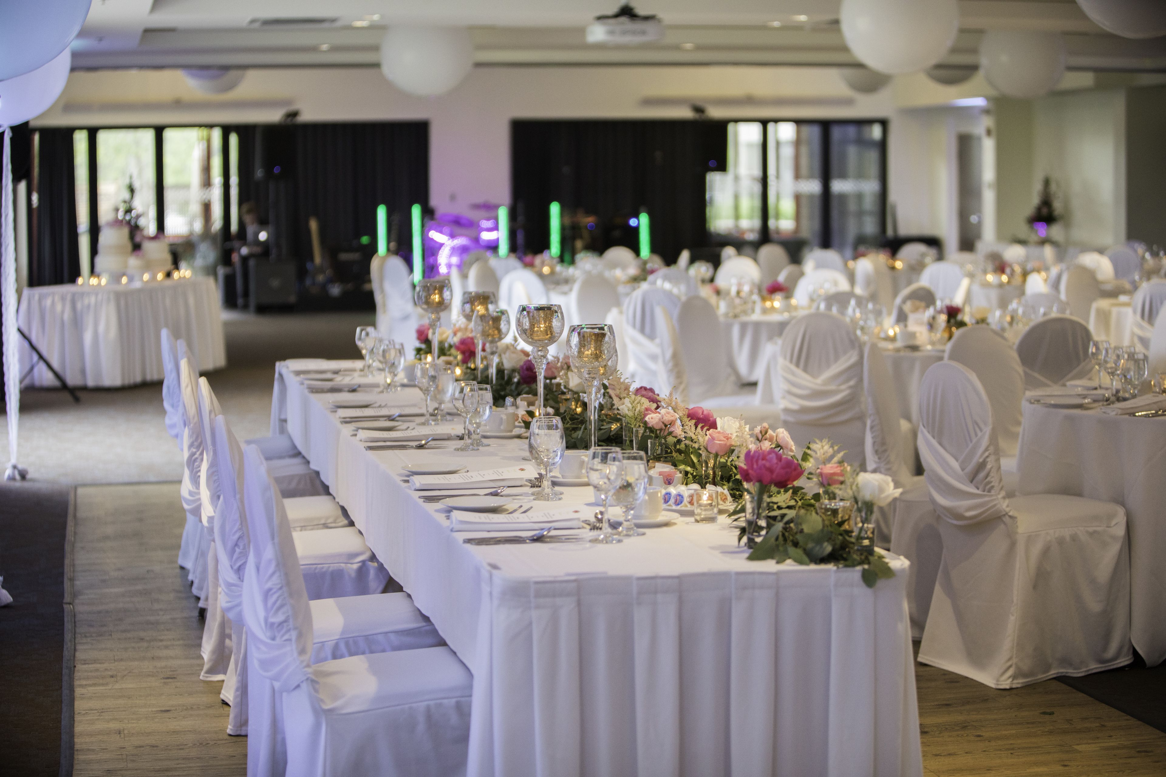 Un salón de banquetes con largas mesas dispuestas para un evento formal, que presenta manteles blancos, sillas con fundas blancas y centros de mesa florales.